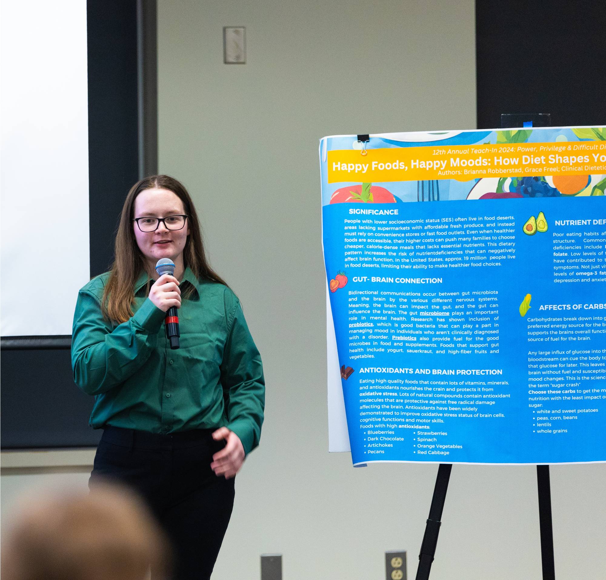 Female student standing in front of a blue poster, with vegetable graphics and text on the poster. Student holding microphone